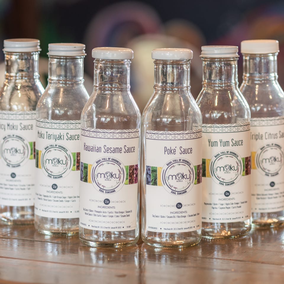 Six bottled Moku Bowls sauces, including teriyaki, sesame, poke, yum yum, and triple citrus, displayed on a wooden counter.
