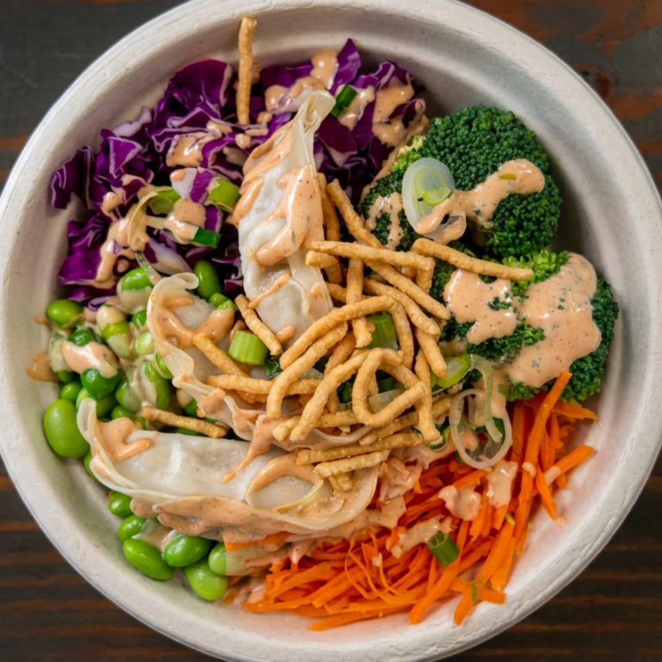 Bowl of vegetable dumplings topped with crispy noodles, broccoli, edamame, red cabbage, shredded carrots, and creamy sauce.