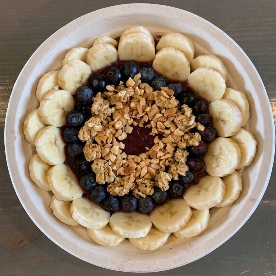 Acai bowl topped with banana slices, blueberries, granola, and chopped nuts on a wooden table.