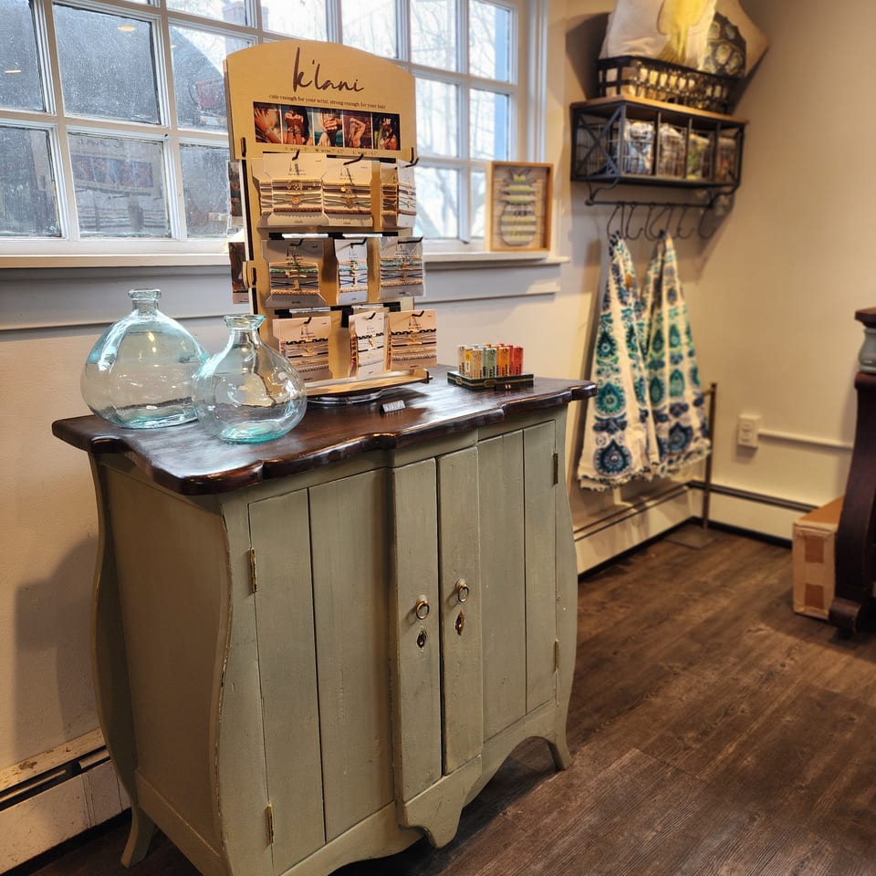 Boutique interior with a vintage cabinet displaying bracelets, glass bottles, patterned garments, and accessories beside large windows.