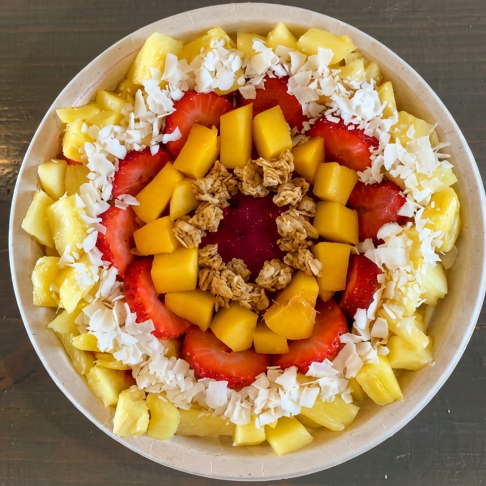 Overhead view of a fruit bowl topped with pineapple, strawberries, mango, shredded coconut, and granola on a wooden table.
