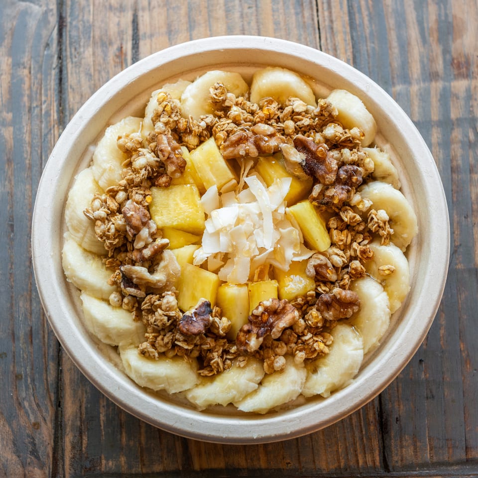 Bowl of sliced bananas, pineapple, granola, walnuts, and coconut flakes drizzled with honey on a rustic wooden table.