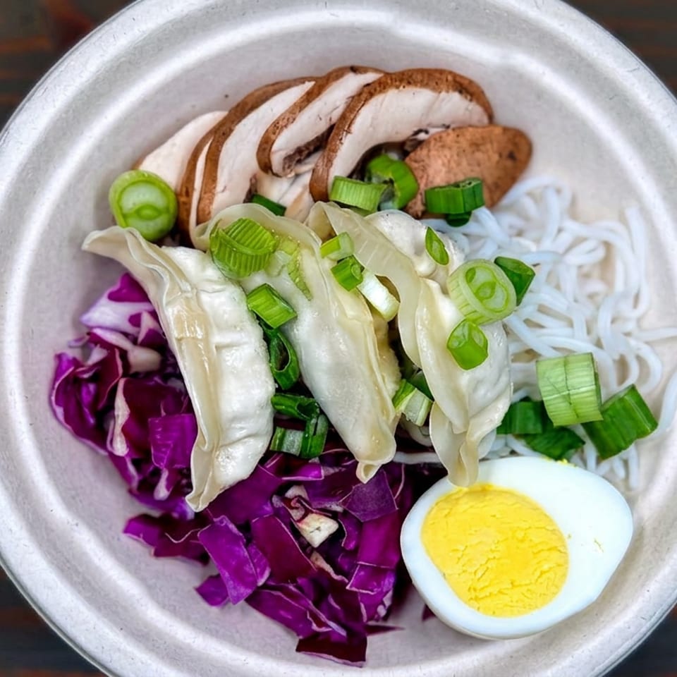 Bowl of noodles topped with dumplings, mushrooms, purple cabbage, sliced scallions, green pepper, and half a boiled egg.