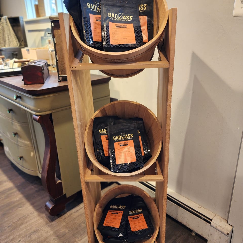 Wooden display rack holds baskets of Bad Ass Coffee Hawaii bags, including coconut rum and vanilla macnut flavors, inside a café.