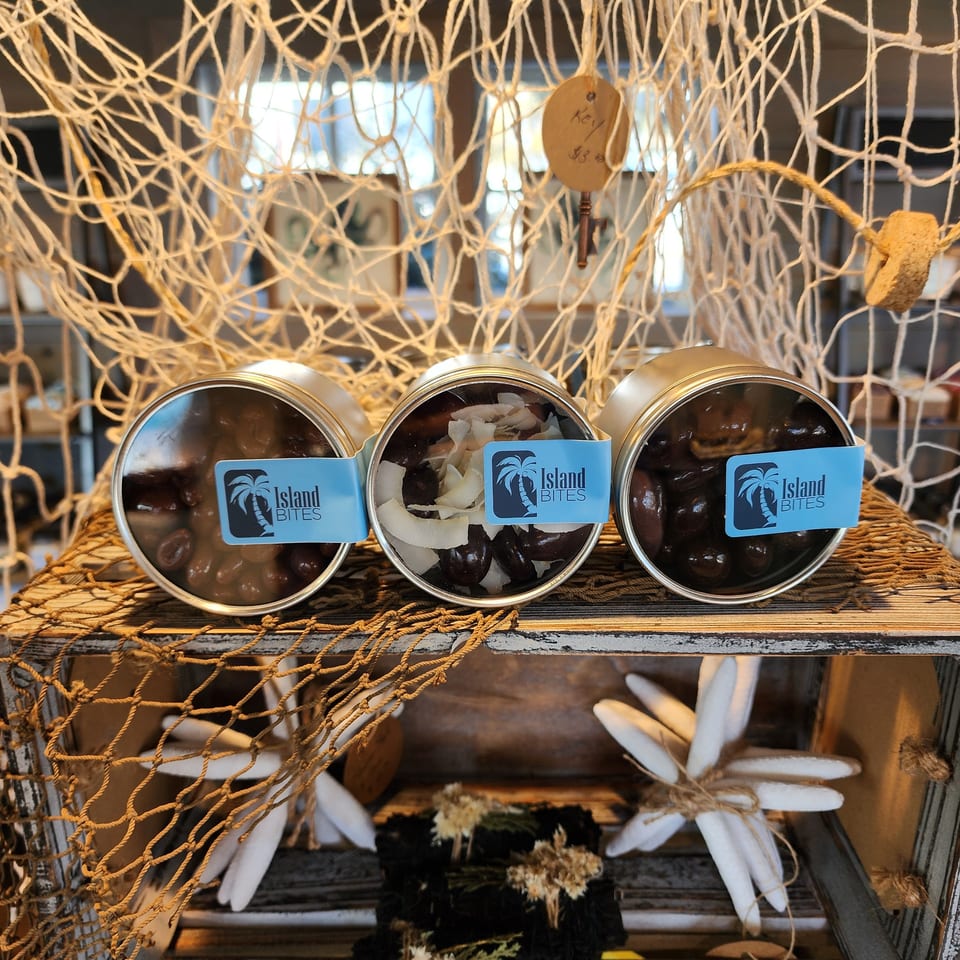 Three open Island Bites tins display dark and white snack pieces on a rustic shelf surrounded by nautical netting and decor.