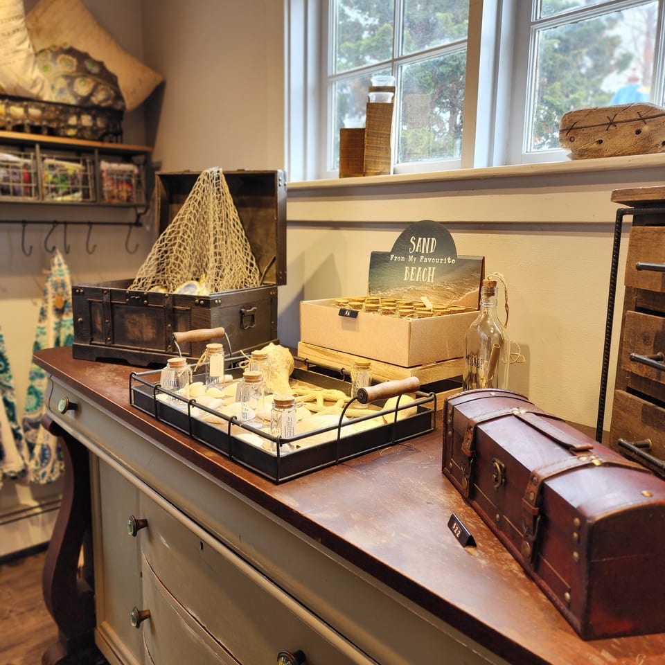 Rustic shop display featuring beach-themed souvenirs, wooden chests, jars, and handcrafted items beside a sunlit window.