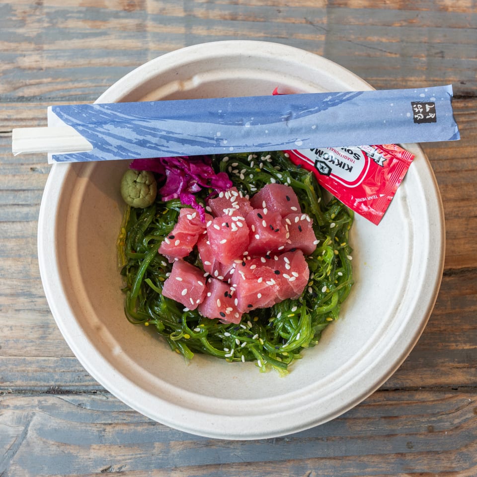Overhead view of a tuna poke bowl with seaweed, sesame seeds, pickled cabbage, wasabi, chopsticks, and soy sauce on a wooden table.