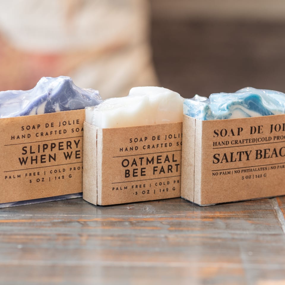 Three handcrafted Soap de Jolie bars in labeled kraft-paper packaging sit on a wooden countertop.