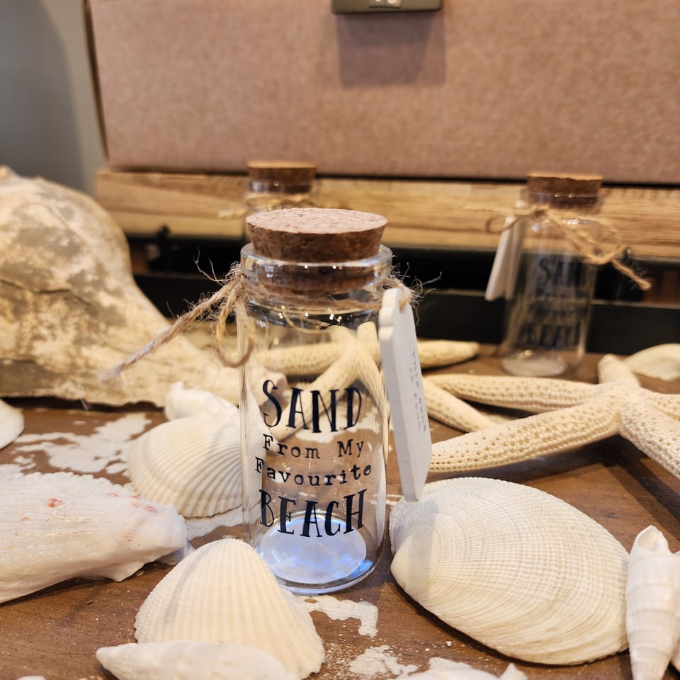 Decorative glass jar labeled “Sand from My Favourite Beach,” surrounded by seashells on a rustic wooden table.