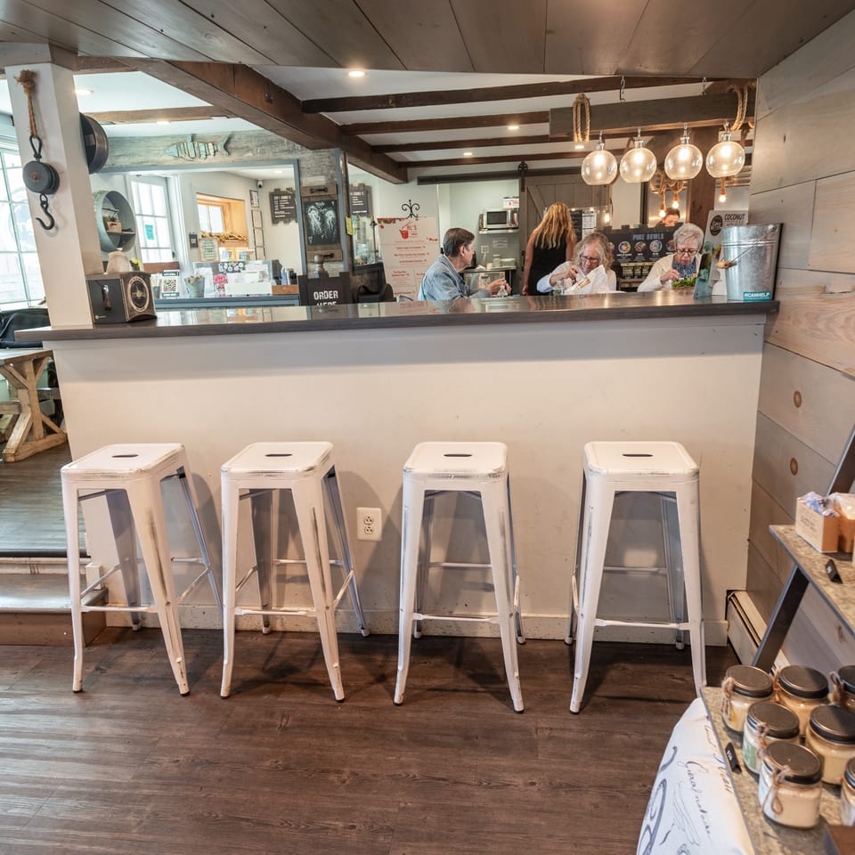 Rustic restaurant counter with white stools, pendant lighting, menu boards, staff, and diners enjoying meals.