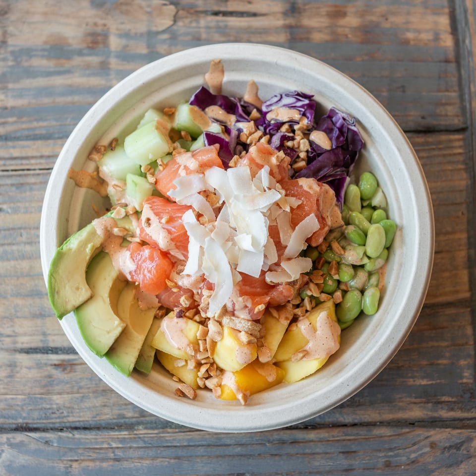 Bowl of salmon poke topped with avocado, edamame, cucumber, cabbage, pineapple, coconut flakes, peanuts, and creamy sauce.