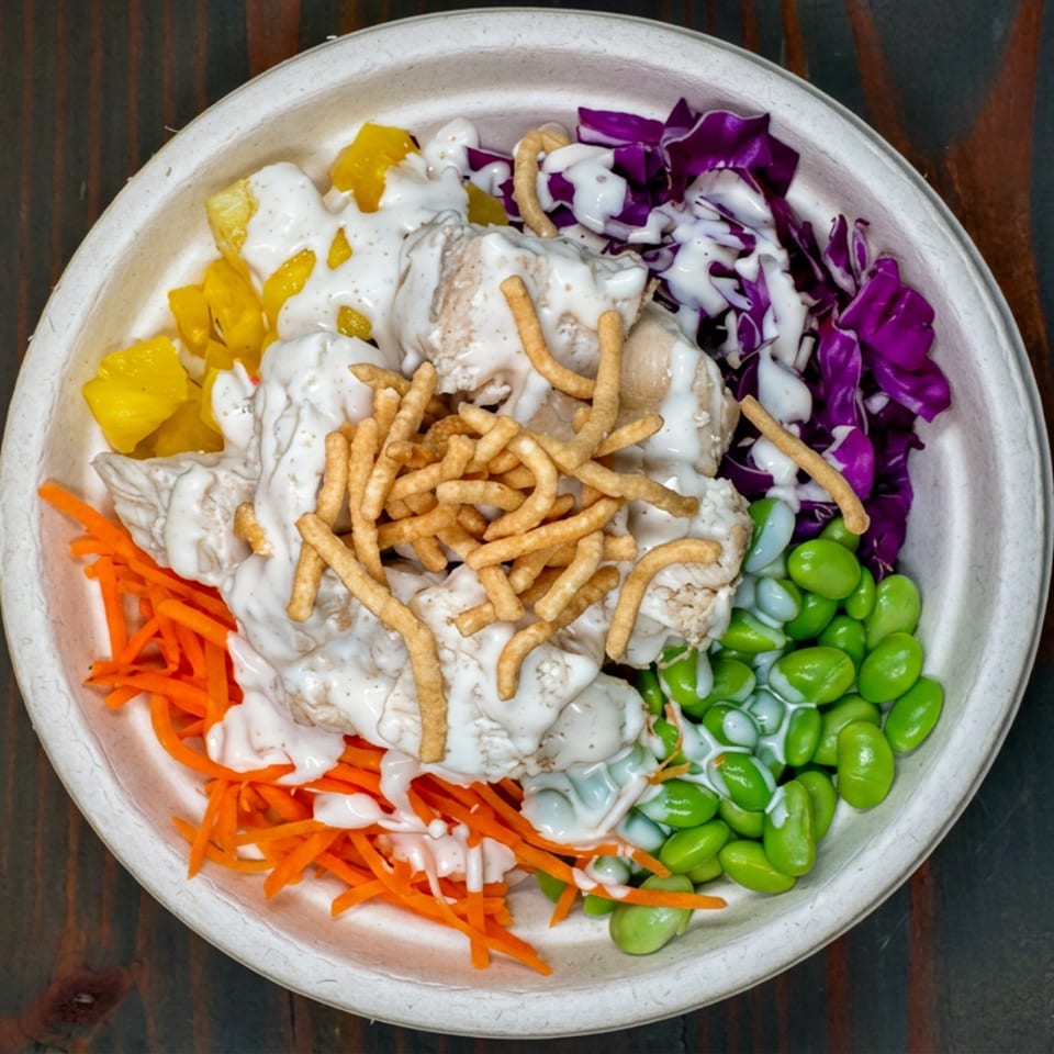Overhead view of a colorful salad bowl with chicken, edamame, cabbage, carrots, mango, creamy dressing, and crispy noodles.