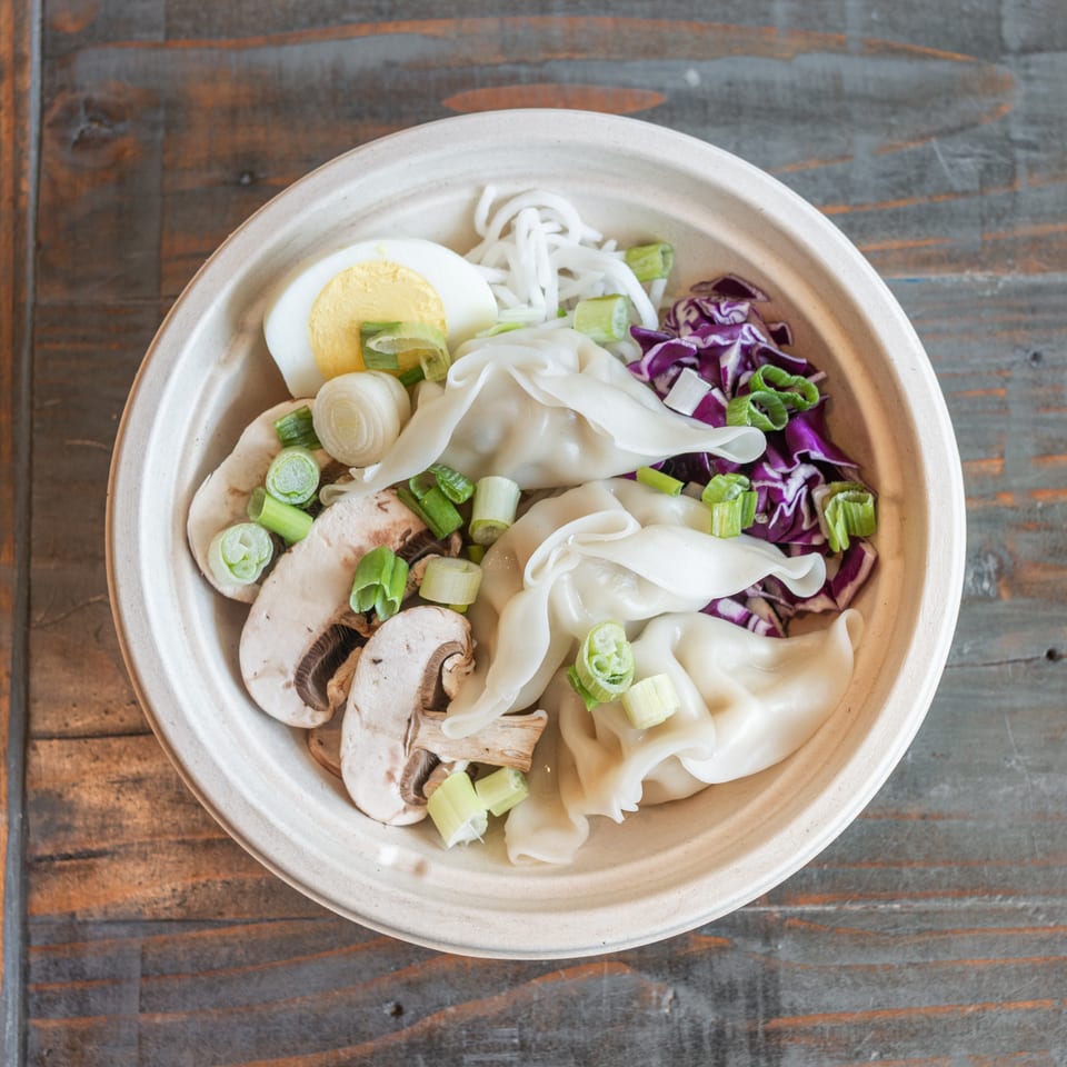 Top-down view of a bowl of dumplings and noodles with mushrooms, cabbage, scallions, and half a boiled egg on a wooden table.