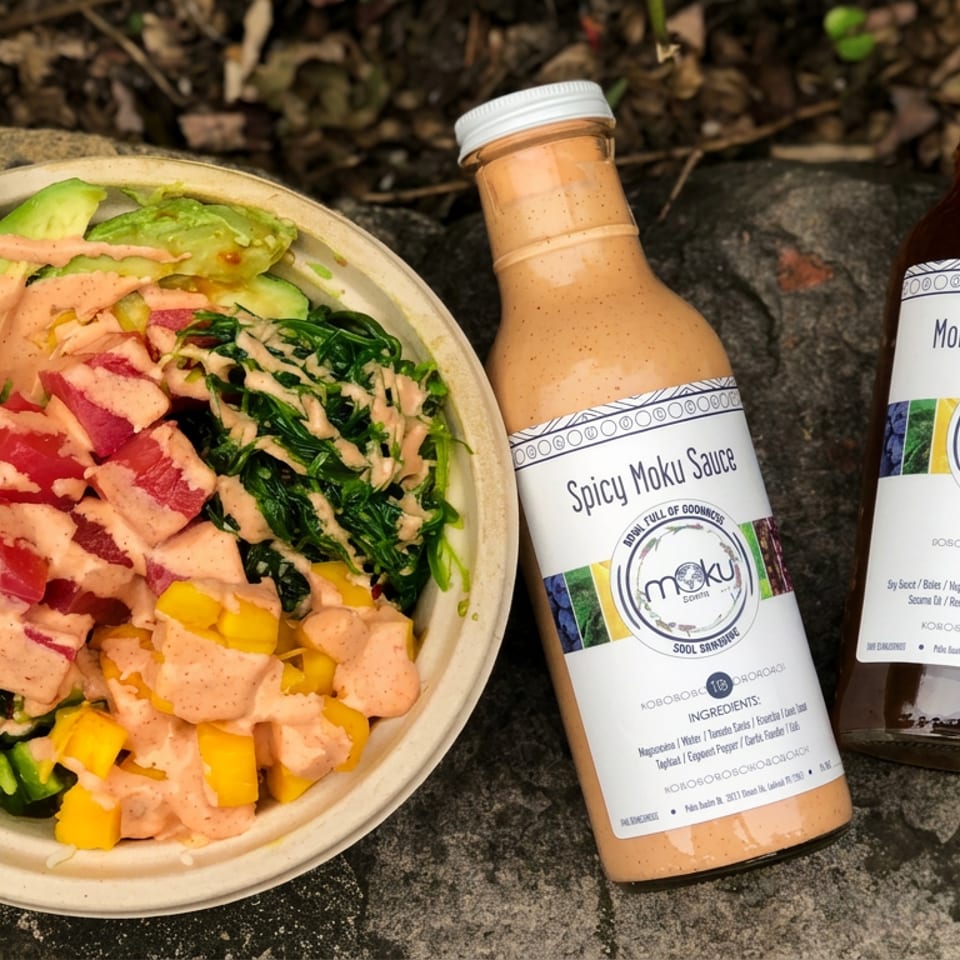 A colorful poke bowl with tuna, avocado, greens, and mango beside bottles of Spicy Moku Sauce and Moku Teriyaki Sauce.